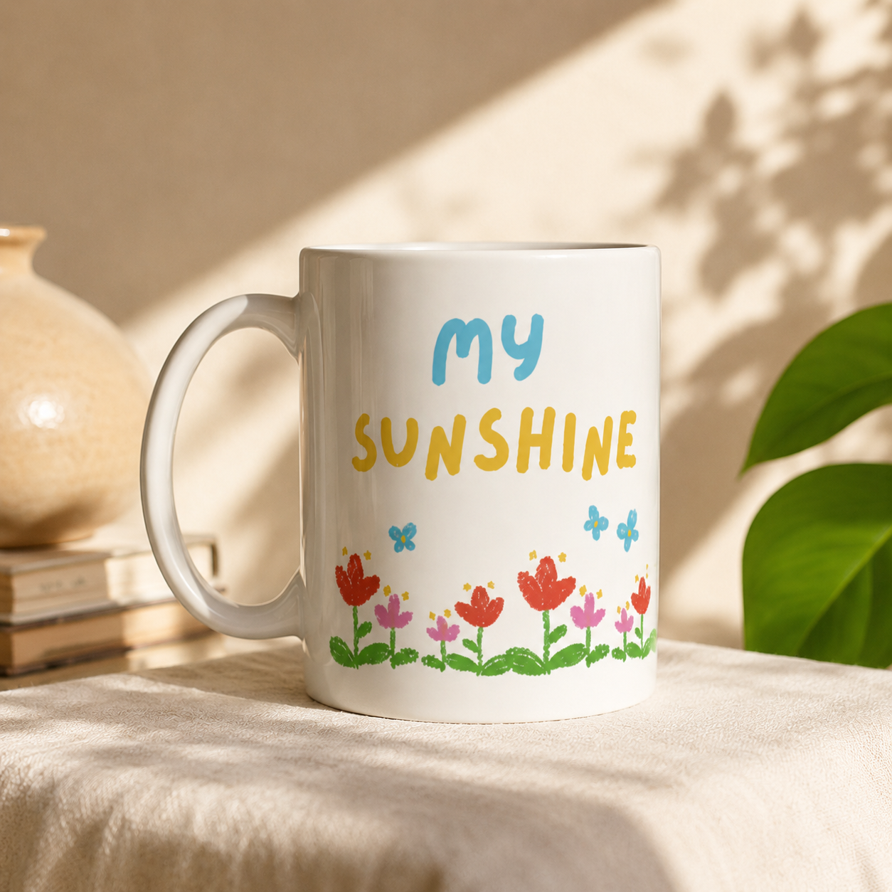 A white ceramic mug with the words 'my SUNSHINE' in blue and yellow, decorated with colorful flowers and butterflies, sitting on a beige fabric surface with warm natural light and shadows.